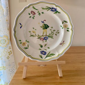 May include: A white ceramic plate with a scalloped edge, featuring a hand-painted central figure of a person in a green jacket and yellow trousers holding a staff, surrounded by colorful floral motifs in blue, yellow, and green.