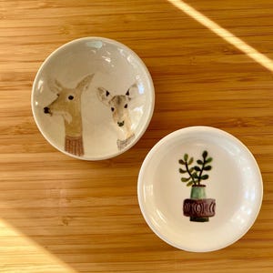 May include: Two small, round ceramic dishes. One dish features a deer illustration, the other a potted plant. The deer dish has two deer heads with brown fur and a beige background. The plant dish has a green plant in a brown pot.