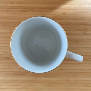 May include: A white teacup with a gold rim and a white handle. The cup is empty and sits on a wooden surface. The cup is viewed from above.