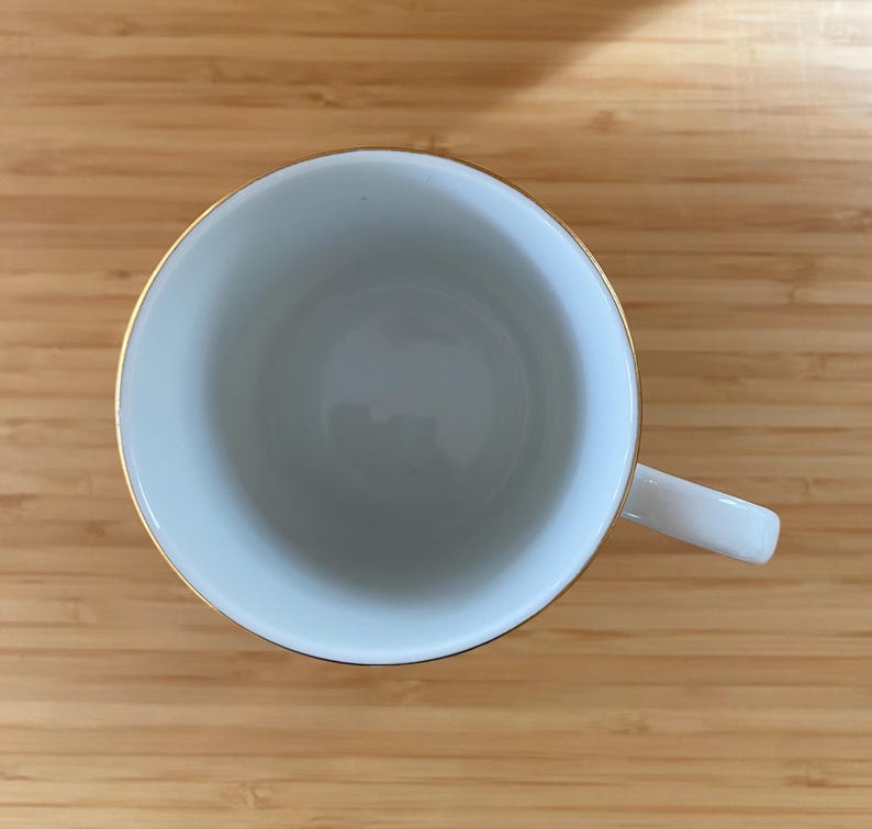 May include: A white teacup with a gold rim and a white handle. The cup is empty and sits on a wooden surface. The cup is viewed from above.