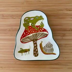 May include: A white ceramic trinket dish with an irregular shape and a dark green rim. The surface features a green frog sitting on a red mushroom with white spots, a brown and green grasshopper, and a brown and white snail with a spiral shell.