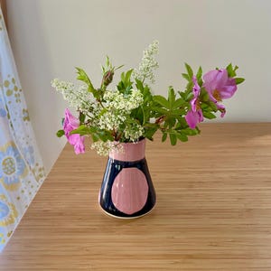 May include: A ceramic vase with a tapered shape, featuring a glossy dark navy blue base with large, rounded light pink accents. The vase is filled with a bouquet of small white clustered flowers and vibrant pink wild rose blossoms with green leaves.