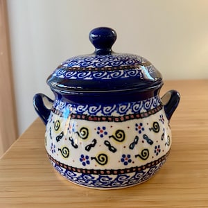 May include: A small, ceramic sugar bowl with a lid. The bowl is white with a blue and black floral pattern and two small handles. The lid and knob are solid blue.