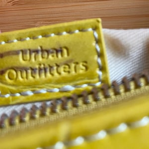 Puede incluir: Primer plano de una etiqueta de cuero amarilla con el texto en relieve "Urban Outfitters". La etiqueta est&aacute; cosida con hilo blanco y est&aacute; unida a una cremallera amarilla. El fondo es una tela beige claro.