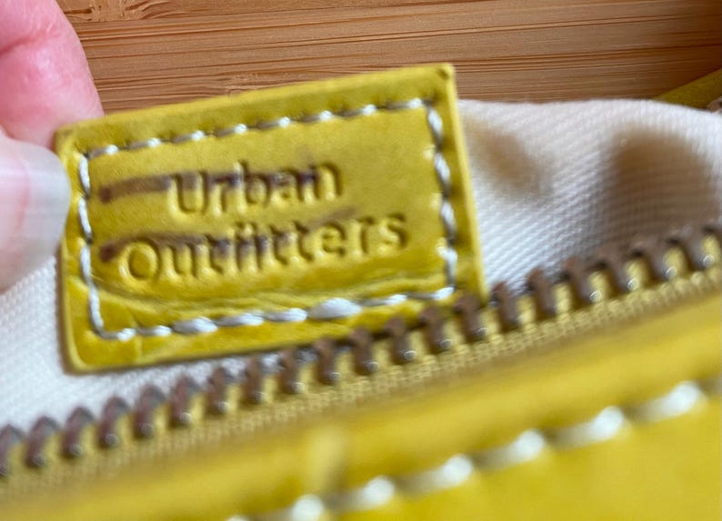 Puede incluir: Primer plano de una etiqueta de cuero amarilla con el texto en relieve "Urban Outfitters". La etiqueta est&aacute; cosida con hilo blanco y est&aacute; unida a una cremallera amarilla. El fondo es una tela beige claro.