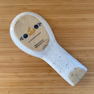May include: White ceramic spoon rest with speckled design. The rest features a light brown speckled area with a chicken illustration and the text "by ANTHROPOLOGIE". Dishwasher and microwave safe.