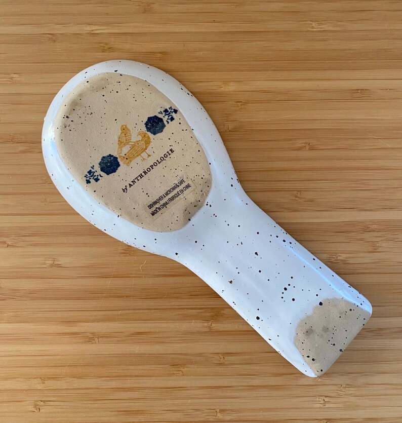 May include: White ceramic spoon rest with speckled design. The rest features a light brown speckled area with a chicken illustration and the text "by ANTHROPOLOGIE". Dishwasher and microwave safe.