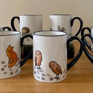 Może przedstawiać: Zestaw białych ceramicznych kubk&oacute;w z czarnymi uchwytami i obw&oacute;dkami. Każdy kubek przedstawia ilustrację leśnego zwierzęcia, w tym lisy, sowy, wiewi&oacute;rki i jeże, z jesiennymi liśćmi. Kubki są ustawione na drewnianej powierzchni.