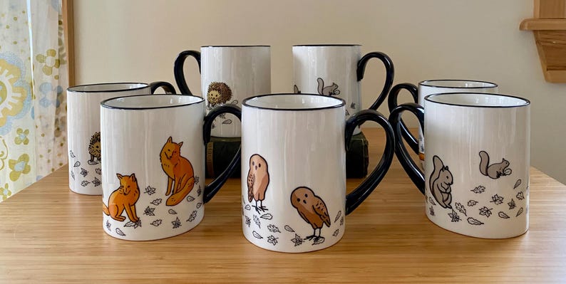 Może przedstawiać: Zestaw białych ceramicznych kubk&oacute;w z czarnymi uchwytami i obw&oacute;dkami. Każdy kubek przedstawia ilustrację leśnego zwierzęcia, w tym lisy, sowy, wiewi&oacute;rki i jeże, z jesiennymi liśćmi. Kubki są ustawione na drewnianej powierzchni.