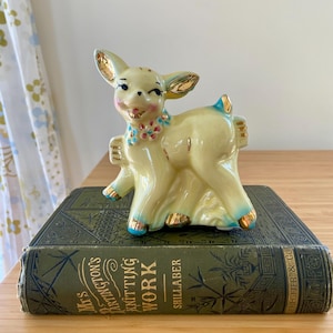May include: A vintage ceramic deer figurine in a light yellow hue with gold accents on the ears and hooves. The deer has a floral collar and is posed on top of a green book with gold lettering. The book's title is "Mrs. Partington's Knitting Work".