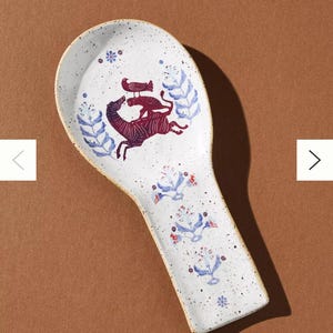 May include: White ceramic spoon rest with a speckled finish and a decorative design. The design features a maroon zebra-like animal with a bird on its back, surrounded by blue floral and leaf motifs.
