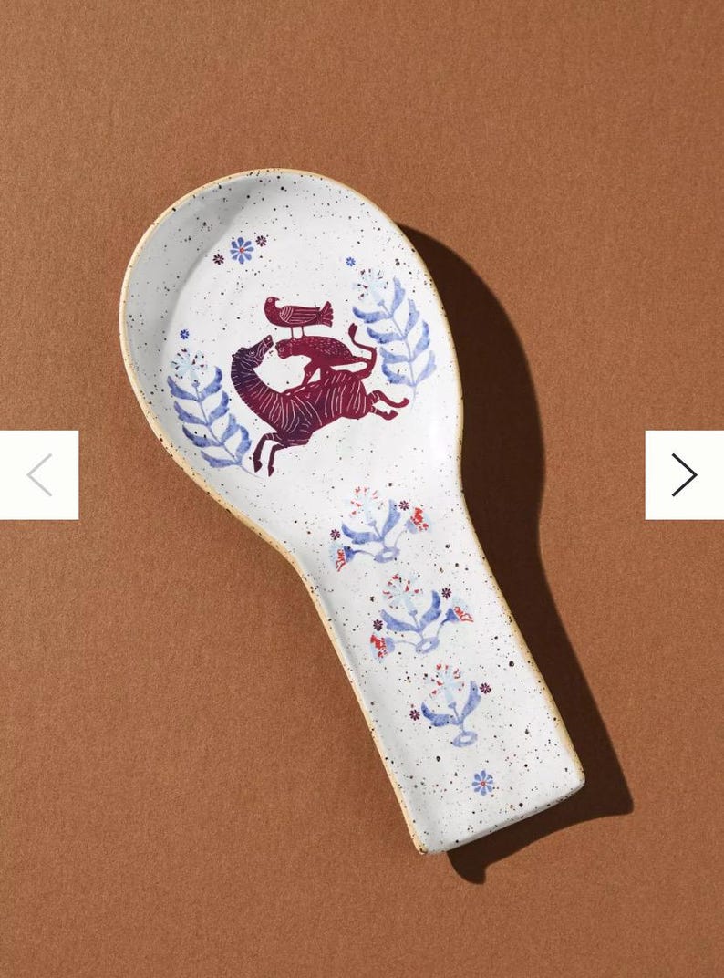 May include: White ceramic spoon rest with a speckled finish and a decorative design. The design features a maroon zebra-like animal with a bird on its back, surrounded by blue floral and leaf motifs.