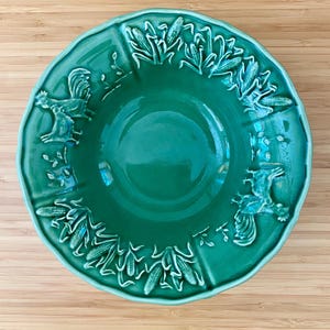 May include: A teal ceramic serving bowl with a scalloped edge. The bowl features embossed corn and rooster designs around the rim. The bowl is set on a light brown wooden surface.