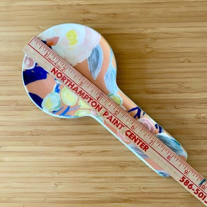 May include: A ceramic spoon rest with a floral design in peach, blue, yellow, and white. A wooden ruler is placed on top, with the text "NORTHAMPTON PAINT CENTER" and measurements in inches.