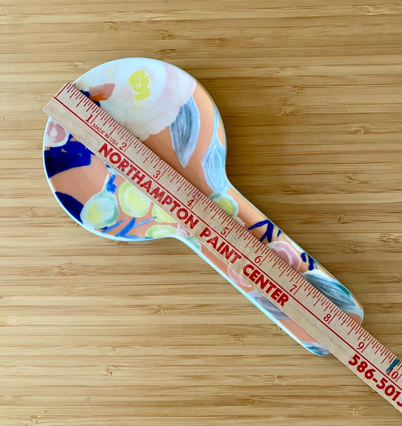 May include: A ceramic spoon rest with a floral design in peach, blue, yellow, and white. A wooden ruler is placed on top, with the text "NORTHAMPTON PAINT CENTER" and measurements in inches.