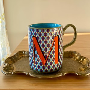 Puede incluir: Taza de cer&aacute;mica decorativa con interior azul, asa verde y un colorido patr&oacute;n geom&eacute;trico. La taza presenta una gran letra "M" naranja y se encuentra sobre una bandeja dorada vintage.