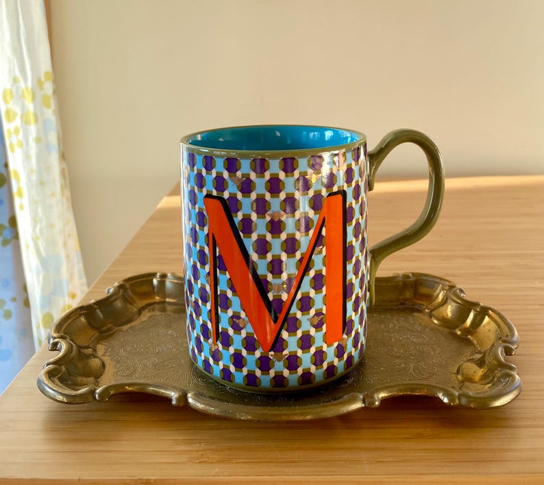 Puede incluir: Taza de cer&aacute;mica decorativa con interior azul, asa verde y un colorido patr&oacute;n geom&eacute;trico. La taza presenta una gran letra "M" naranja y se encuentra sobre una bandeja dorada vintage.