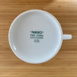 May include: White ceramic teacup with a handle, viewed from the bottom. The text "NIKKO FINE CHINA GOLD FILIGREE PATRA THAILAND" is printed in green. The cup is on a light brown wooden surface.