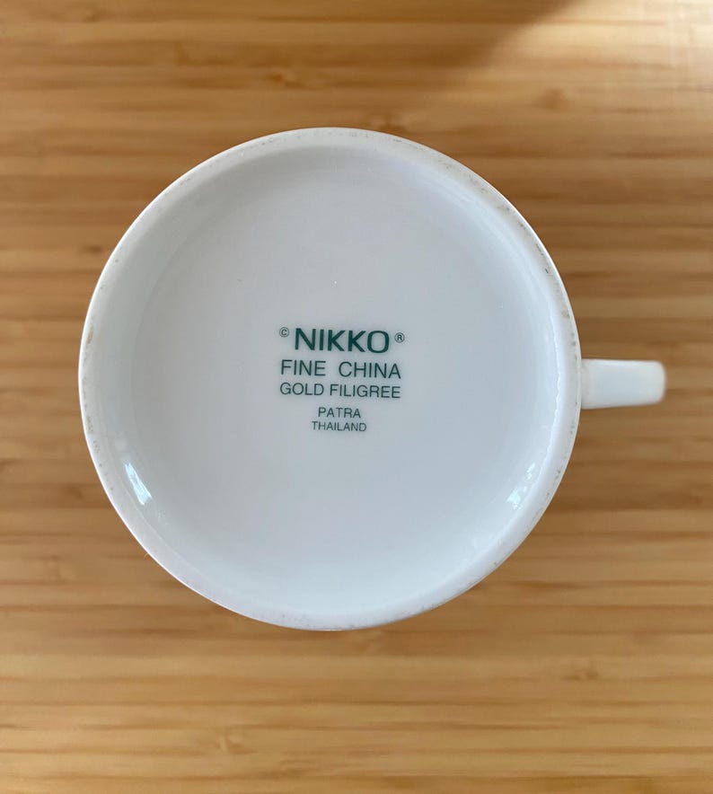 May include: White ceramic teacup with a handle, viewed from the bottom. The text "NIKKO FINE CHINA GOLD FILIGREE PATRA THAILAND" is printed in green. The cup is on a light brown wooden surface.