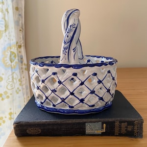 May include: A white and blue ceramic basket with a lattice design and a handle. The basket is decorated with blue accents and sits atop a dark blue book. The handle has a twisted design with floral patterns.