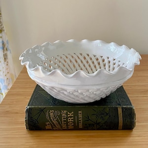 May include: A white ceramic bowl with a lattice-style woven design and a ruffled, scalloped edge. The bowl features a glossy finish and sits atop a dark green hardcover book titled Mrs. Partington's Knitting Work by Shillaber.