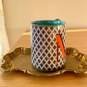 Puede incluir: Una taza de cer&aacute;mica estampada con interior turquesa y una letra "N" naranja en el lateral, apoyada sobre una bandeja dorada. La taza presenta un dise&ntilde;o geom&eacute;trico en morado, azul y dorado. La bandeja tiene un borde decorativo festoneado.