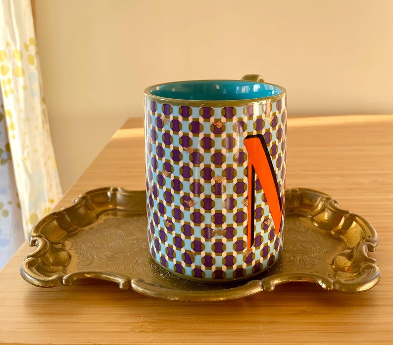 Puede incluir: Una taza de cer&aacute;mica estampada con interior turquesa y una letra "N" naranja en el lateral, apoyada sobre una bandeja dorada. La taza presenta un dise&ntilde;o geom&eacute;trico en morado, azul y dorado. La bandeja tiene un borde decorativo festoneado.