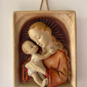 May include: A rectangular relief sculpture depicting a mother and child. The mother, with flowing blonde hair and a red robe, cradles the baby. The background features a halo and a warm color palette, with a beige frame and a hanging cord.