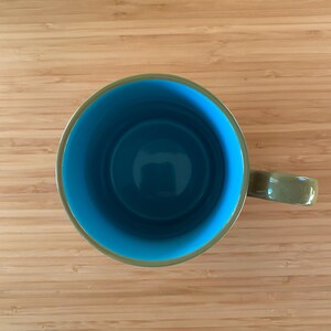 Puede incluir: Vista a&eacute;rea de una taza de cer&aacute;mica con interior turquesa y exterior verde. El asa de la taza tambi&eacute;n es verde. La taza est&aacute; sobre una superficie de madera marr&oacute;n claro.