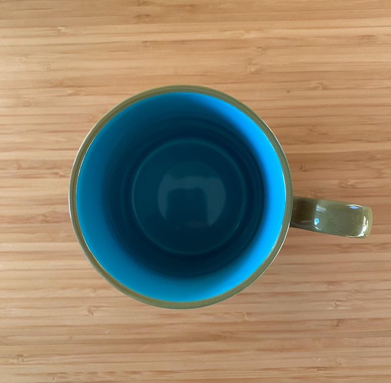 Puede incluir: Vista a&eacute;rea de una taza de cer&aacute;mica con interior turquesa y exterior verde. El asa de la taza tambi&eacute;n es verde. La taza est&aacute; sobre una superficie de madera marr&oacute;n claro.
