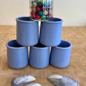 May include: A clear glass jar filled with colorful wooden beads sits atop a pyramid of five light blue ceramic cups. Three seashells are arranged at the base, offering a contrast in texture and color.