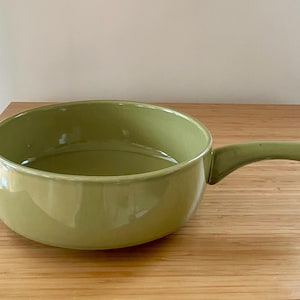 May include: A light green ceramic saucepan with a single handle. The pan is round with a wide opening and sits on a wooden surface. The handle curves slightly downward.