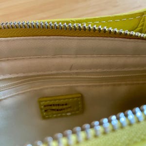 Puede incluir: Primer plano del interior de un bolso amarillo con cremallera plateada. El interior es de color crema claro con una peque&ntilde;a etiqueta rectangular amarilla. El bolso est&aacute; abierto, revelando el forro interior y un peque&ntilde;o bolsillo.