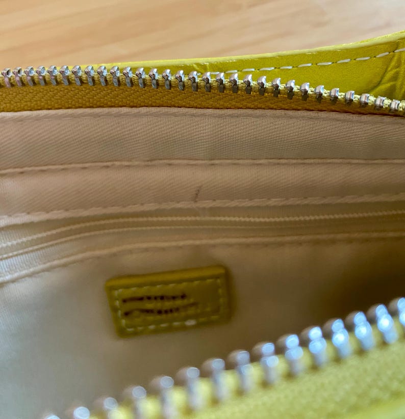 Puede incluir: Primer plano del interior de un bolso amarillo con cremallera plateada. El interior es de color crema claro con una peque&ntilde;a etiqueta rectangular amarilla. El bolso est&aacute; abierto, revelando el forro interior y un peque&ntilde;o bolsillo.
