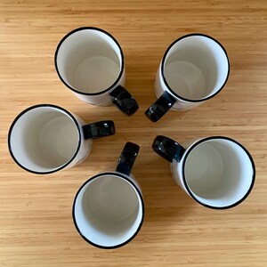 Może przedstawiać: Pięć białych ceramicznych kubk&oacute;w z czarnymi obw&oacute;dkami i uchwytami ułożonych w okrąg na drewnianej powierzchni. Kubki mają prosty, nowoczesny design, odpowiedni do kawy lub herbaty.