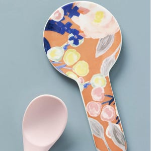 May include: A ceramic spoon rest with a floral design in orange, pink, blue, and yellow. The spoon rest has a handle and a round head. A pink silicone spoon with a wooden handle is next to the spoon rest.