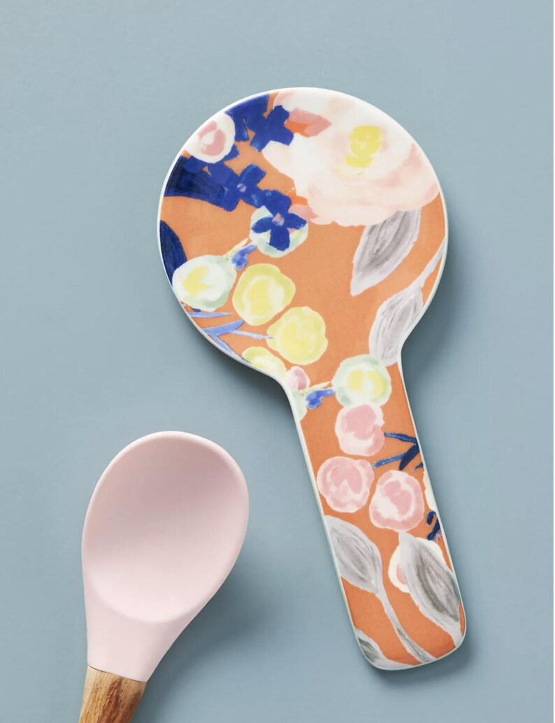 May include: A ceramic spoon rest with a floral design in orange, pink, blue, and yellow. The spoon rest has a handle and a round head. A pink silicone spoon with a wooden handle is next to the spoon rest.