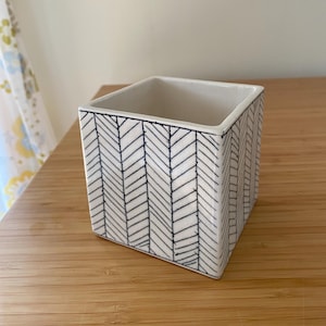 May include: A square ceramic planter featuring a white base with a hand-drawn black herringbone pattern. The vessel has a clean, open top and straight vertical sides, showcasing a consistent geometric design across its exterior surfaces.
