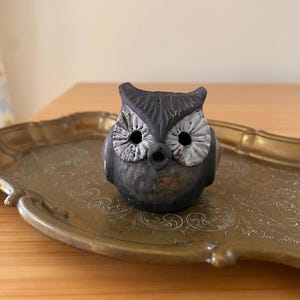 May include: A small, dark gray owl figurine with white accents on the eyes and a textured surface. The owl is perched on a gold-colored, ornate tray with a detailed pattern.