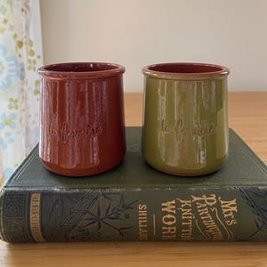 May include: Two ceramic jars, one reddish-brown and one olive green, with the text "la fermière" embossed on the front. The jars are cylindrical with a slightly flared rim. They sit on a vintage book.