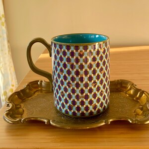 Puede incluir: Una taza de cer&aacute;mica estampada con interior turquesa y asa dorada descansa sobre una bandeja decorativa dorada. La taza presenta un dise&ntilde;o geom&eacute;trico repetitivo en morado, dorado y azul claro. La bandeja tiene un borde ornamentado y festoneado.