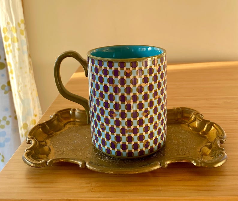 Puede incluir: Una taza de cer&aacute;mica estampada con interior turquesa y asa dorada descansa sobre una bandeja decorativa dorada. La taza presenta un dise&ntilde;o geom&eacute;trico repetitivo en morado, dorado y azul claro. La bandeja tiene un borde ornamentado y festoneado.