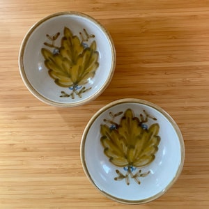 May include: Two small, round ceramic bowls with a white interior and a tan rim. Each bowl features a hand-painted design of a yellow-green leaf with blue accents. The bowls are set on a light brown wooden surface.