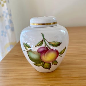 May include: A white porcelain jar with a lid featuring a gold band. The body of the jar is decorated with a hand-painted illustration of two plums, one green and one purple-yellow, surrounded by green leaves on a branch against a white background.