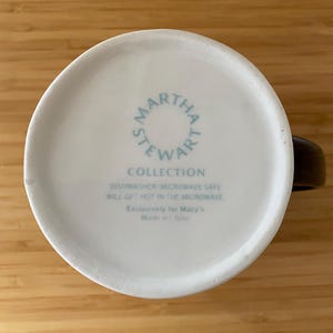 Może przedstawiać: Baza białego ceramicznego kubka z napisem "MARTHA STEWART COLLECTION" w okrągłym wzorze. Dodatkowy tekst wskazuje, że kubek nadaje się do zmywarki i kuchenki mikrofalowej. Kubek ma brązowy uchwyt.