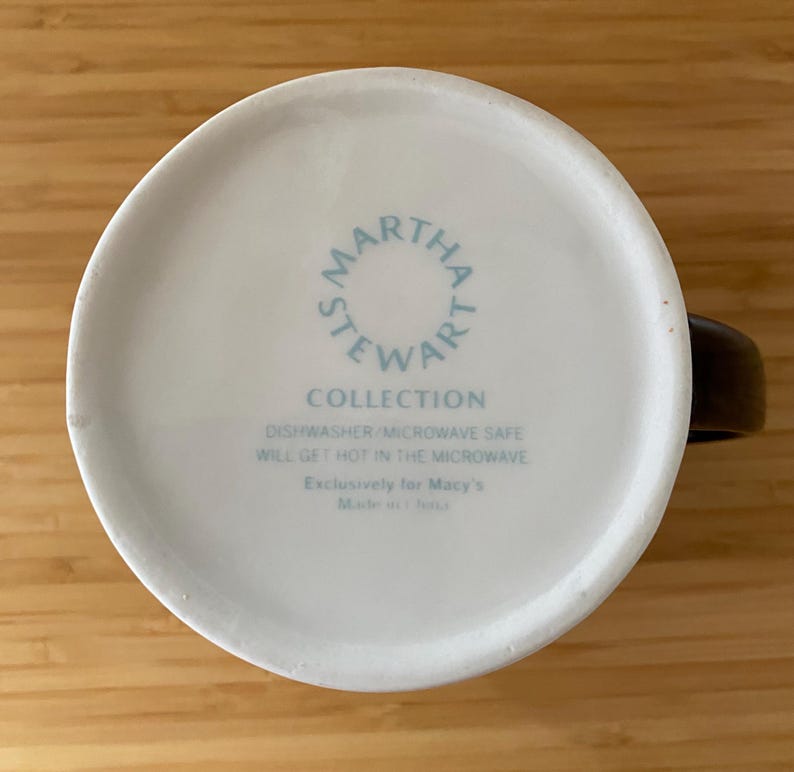 Może przedstawiać: Baza białego ceramicznego kubka z napisem "MARTHA STEWART COLLECTION" w okrągłym wzorze. Dodatkowy tekst wskazuje, że kubek nadaje się do zmywarki i kuchenki mikrofalowej. Kubek ma brązowy uchwyt.