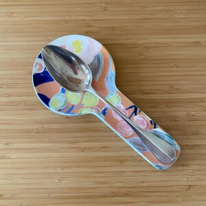 May include: A silver spoon rests on a floral patterned spoon rest. The spoon rest has a peach background with blue, yellow, and pink floral designs. The spoon is made of shiny silver metal.