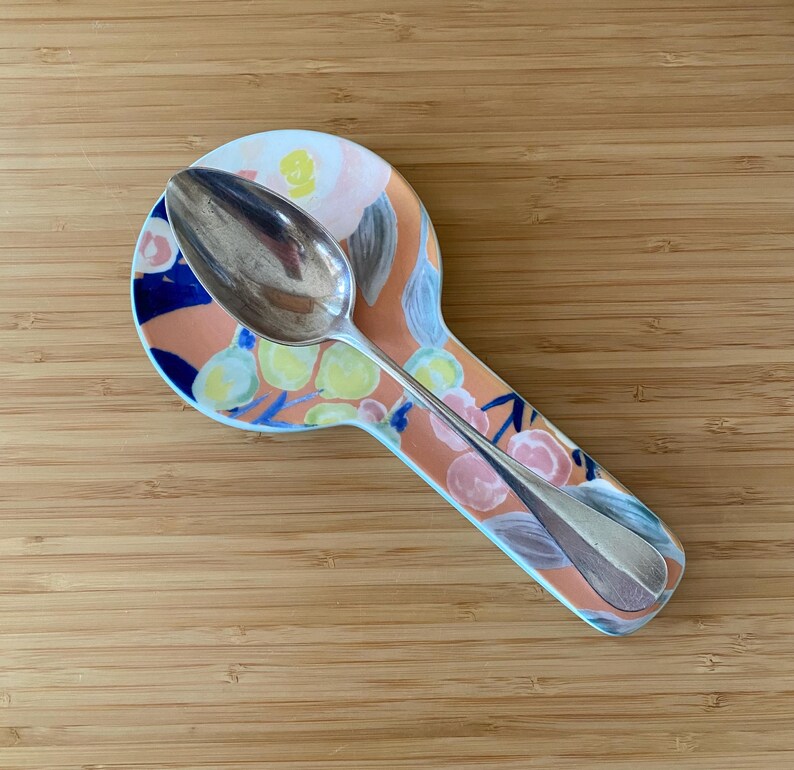 May include: A silver spoon rests on a floral patterned spoon rest. The spoon rest has a peach background with blue, yellow, and pink floral designs. The spoon is made of shiny silver metal.