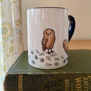 Może przedstawiać: Biały ceramiczny kubek z brązową ilustracją sowy i granatowym uchwytem. Sowa otoczona jest wzorami liści. Obręcz i uchwyt kubka są granatowe.
