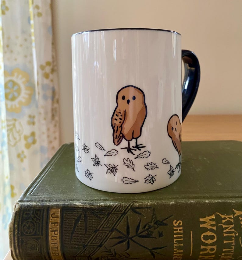 Może przedstawiać: Biały ceramiczny kubek z brązową ilustracją sowy i granatowym uchwytem. Sowa otoczona jest wzorami liści. Obręcz i uchwyt kubka są granatowe.